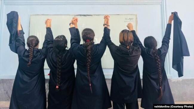 Female students stand in a line without hijab facing a whiteboard in a school in Iran.