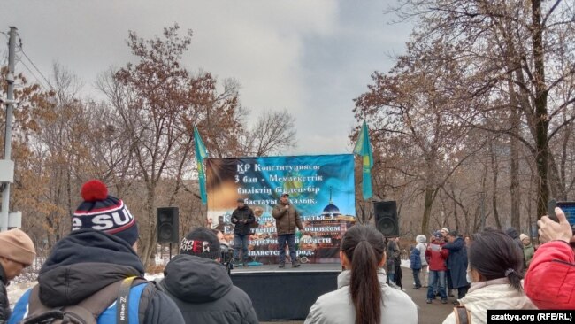 Two men stand on a small stage and speak to an audience.