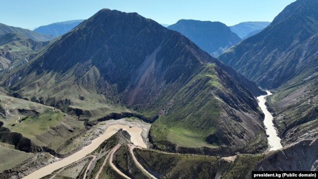 Construction of the Kambar-Ata 1 hydro power plant on June 8, 2022, taking place between giant, green mountains. The river winding between them runs muddy and brown.