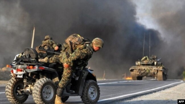 A soldiers hops off an ATV vehicle. Black smoke and fire rise in the background.