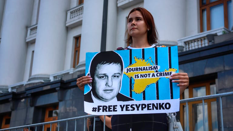 A woman holds a blue and yellow poster with an image of Vladyslav Yesypenko. The text of the posters reads: "Journalism is not a crime. #Free Yesypenko."