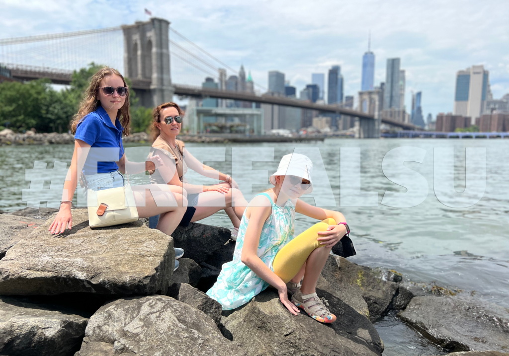 An image of Alsu Kurmasheva and her two daughters sitting on rocks by a river in New York. Alsu is currently unjustly detained in Russia after she was accused of violating Russia's law on foreign agents.